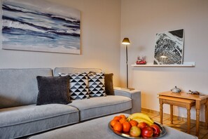 Ein modernes Wohnzimmer mit einem grauen Sofa, gemusterten Kissen, einer Obstschale auf einem Tisch, einer Stehlampe und Kunstwerken an den Wänden.