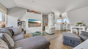Uma sala de estar moderna com uma mesa de centro de vidro, sofás cinzentos, uma poltrona branca e um espaço de jantar com mesa e cadeiras brancas.