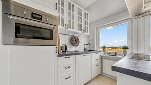 Cozinha com armários brancos, forno de aço inoxidável, janela com vista e bancada com chaleira.