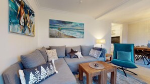 Una sala moderna con un sofá gris, una mesa de centro de madera y un sillón color turquesa. En la pared, hay un cuadro con una escena de playa.