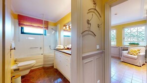 A bathroom with a toilet, bathtub, and shower. There is a sink with a mirror above it. The room has yellow walls and a window with a view of greenery outside. - Luv Biberhüs - Großzügige Wohnung im Landwirtschaftlich Geprägten Archsum mit Gartenteil (Sylt)