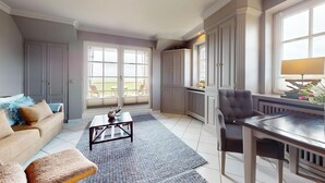 Un salon comprenant un canapé, une table basse et une table à manger avec des chaises. De grandes fenêtres offrent une vue sur l’extérieur.