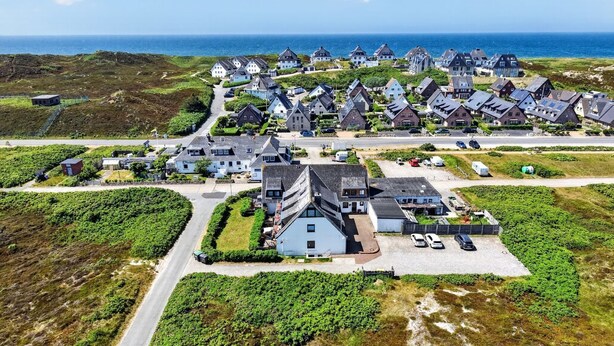 An aerial view of a coastal village with traditional houses, a central building with a sloped roof, and a road leading to the sea. - Fischer - Praktisches Appartement in Strandnaher Lage Inmitten Einer Heidelandschaft (Hörnum (Sylt))