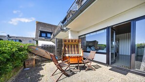 A patio with a table and chairs, a surfboard, and a sliding glass door to a balcony. - Fischer - Praktisches Appartement in Strandnaher Lage Inmitten Einer Heidelandschaft (Hörnum (Sylt))