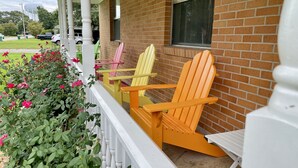 A porch with a brick wall, a white railing, and three Adirondack chairs in yellow, pink, and orange.