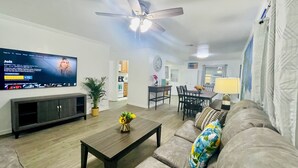 A living room with a flat-screen TV, a ceiling fan, a dining area with a table and chairs, and a sofa with a floral cushion.