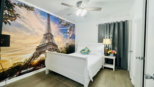 A hotel room with a bed, a nightstand with a lamp and flowers, a ceiling fan, and a mural of the Eiffel Tower.
