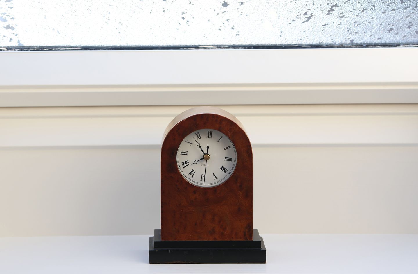 Une horloge en bois, avec des chiffres romains sur un socle noir, posée sur une surface blanche devant un mur blanc, avec une fenêtre au-dessus.