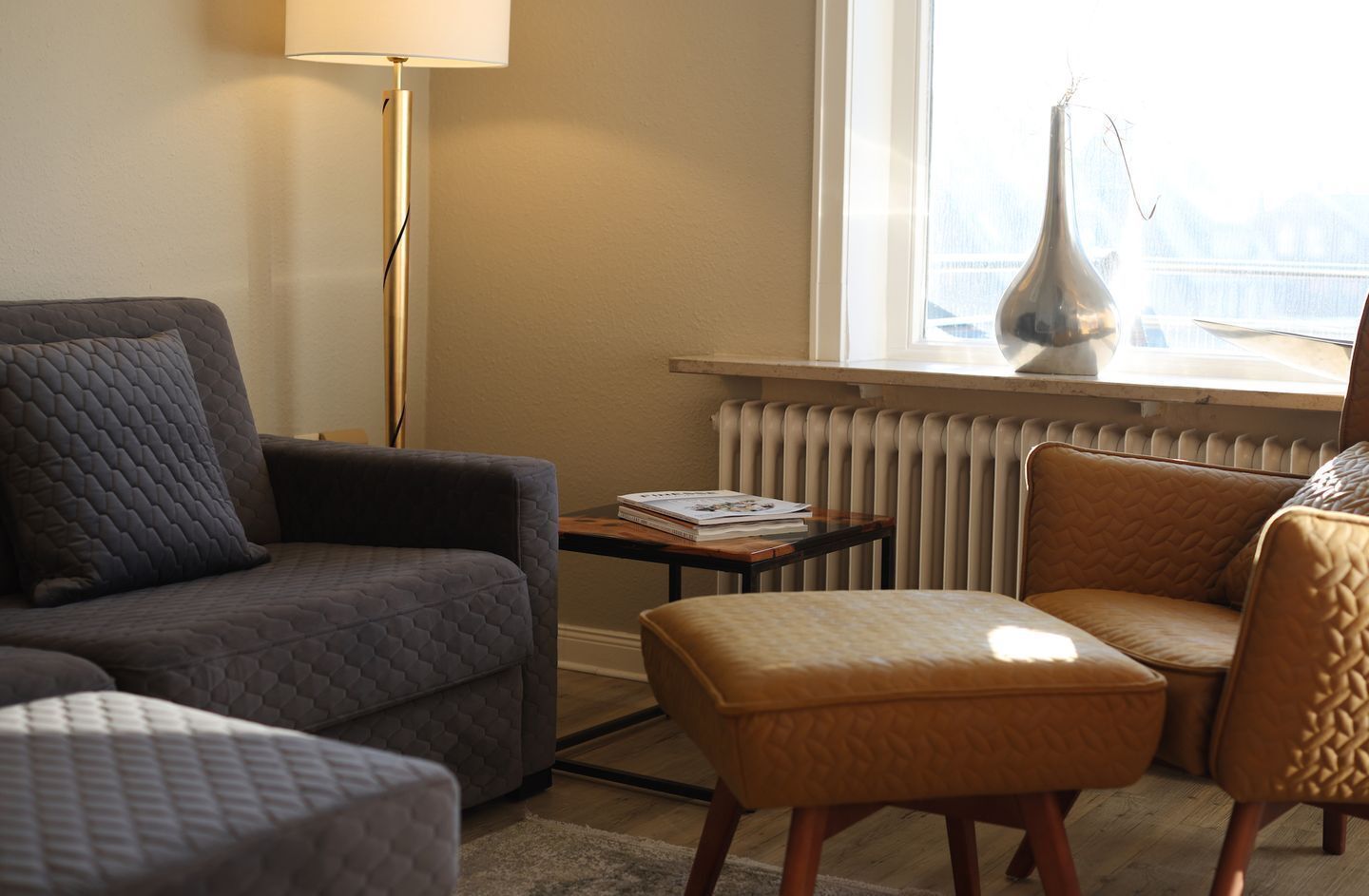 A living room with a grey sofa, a small table with books, a brown armchair, and a radiator.