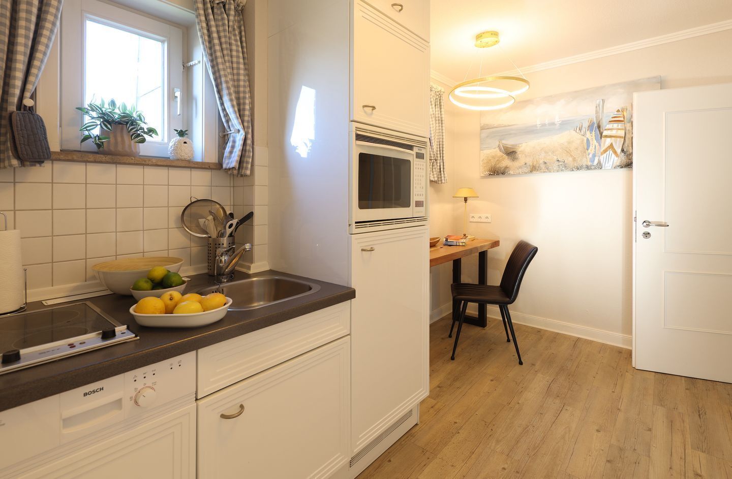 A kitchen with a Bosch dishwasher, a microwave, and a sink with lemons. There is a dining table with a chair and a lamp.