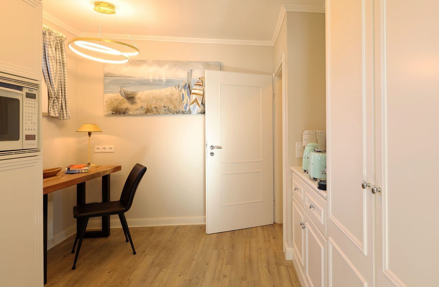 Et kompakt køkken med en mikrobølgeovn, et bord med en stol, en hængende lampe og en dør til et badeværelse med toilet og skab.