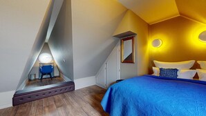 Un dormitorio con techo inclinado, una cama con ropa de cama azul, un espejo y una pequeña mesa con una silla.