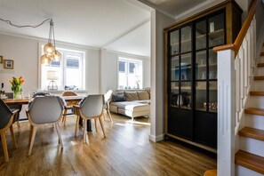 Amplia sala de estar con una mesa de comedor de madera, sillas y un sofá. Se observa un mueble negro con puertas de cristal, y una escalera conduce a otro nivel.