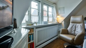 Un dormitorio acogedor en el ático, con un ventanal grande, un radiador, una estantería, un sillón de cuero y un televisor.