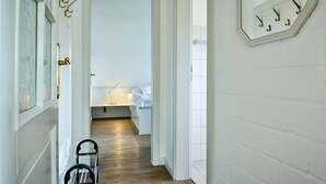 Un pasillo con una cama, un escritorio y una silla. Hay ganchos en la pared y una puerta con manija de bronce.