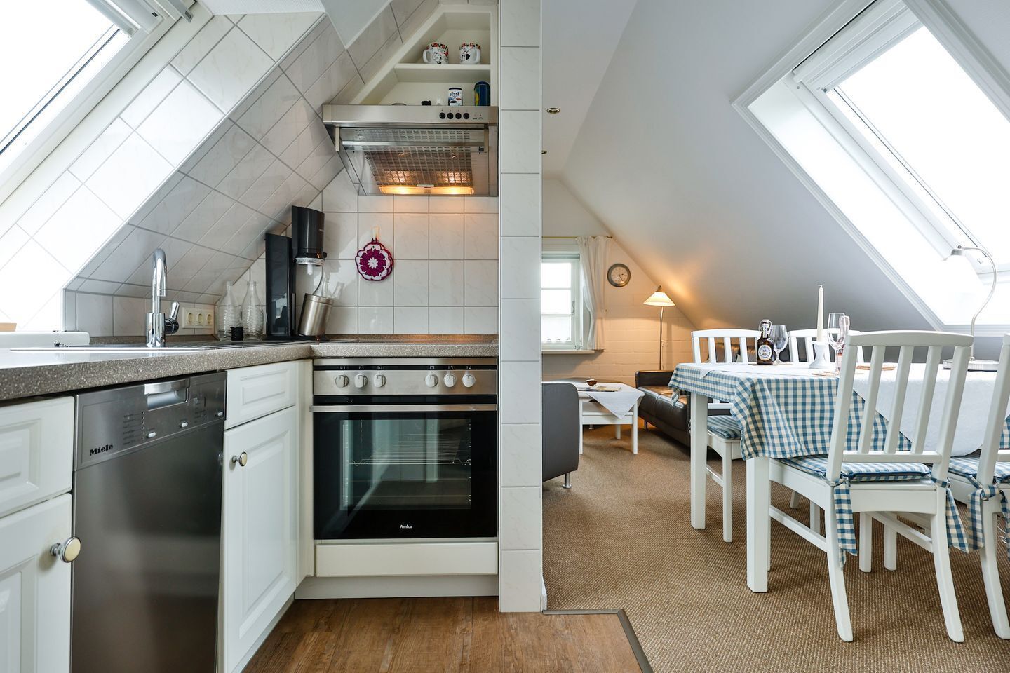 A compact kitchen with a dishwasher, oven, and a small dining area with a checkered tablecloth and white chairs.