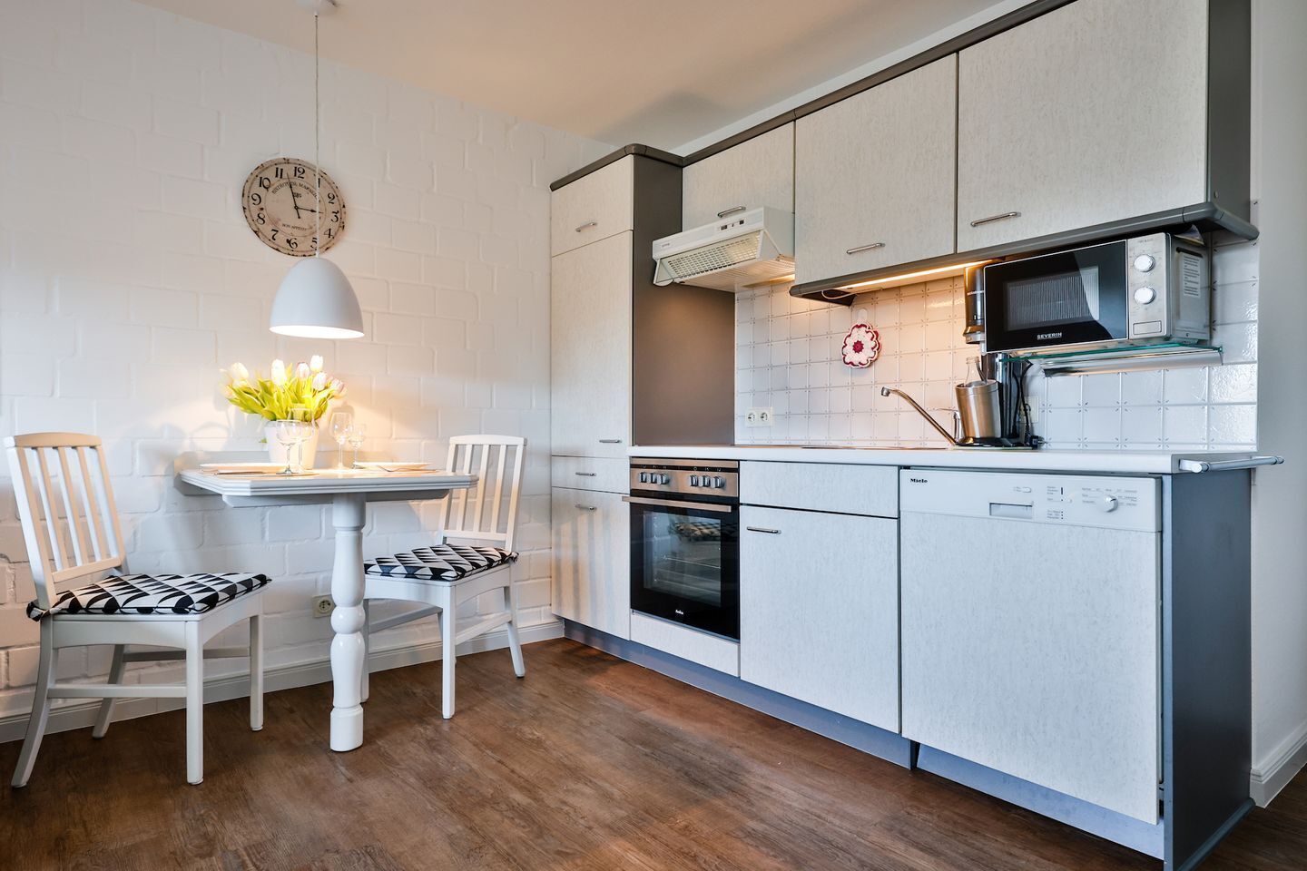 Une cuisine compacte avec des meubles blancs, un four encastré, un four à micro-ondes et un petit coin repas comprenant deux chaises et une table.