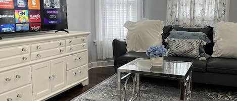 Ett vardagsrum med en platt-TV som visar olika kanaler, en vit byrå, ett glasbord, en mörk soffa och en mönstrad matta.