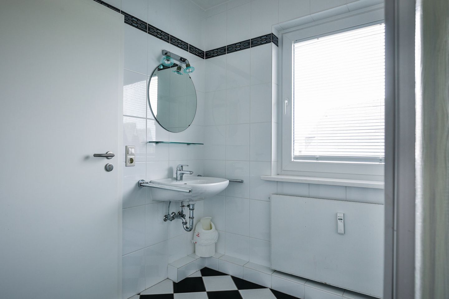 Une salle de bain carrelée de carreaux blancs, avec un lavabo doté d’une étagère en verre, un miroir et une fenêtre avec des stores.