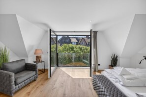 Ein modernes Schlafzimmer mit einem großen Schiebeglastür, die auf einen Balkon führt, einem grauen Sofa, einem Nachttisch mit einer Lampe und einem Bett mit ordentlich gebetteter weißer Bettwäsche.