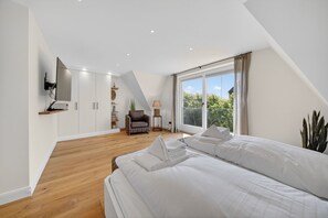A bedroom with a large bed, a chair, a TV mounted on the wall, and a window with curtains.