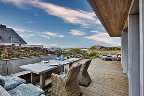 En træterrasse med et bord og stole, med udsigt over en strand og et hus.