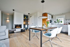 En moderne stue med pejs, et spiseområde med et træbord og hvide stole, og et køkken med hvide skabe og et vindue.