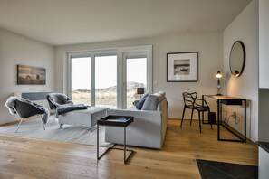 En moderne stue med en grå sofa, to lænestole, et sofabord og et skrivebord med en lampe. Store vinduer med udsigt til det fri.