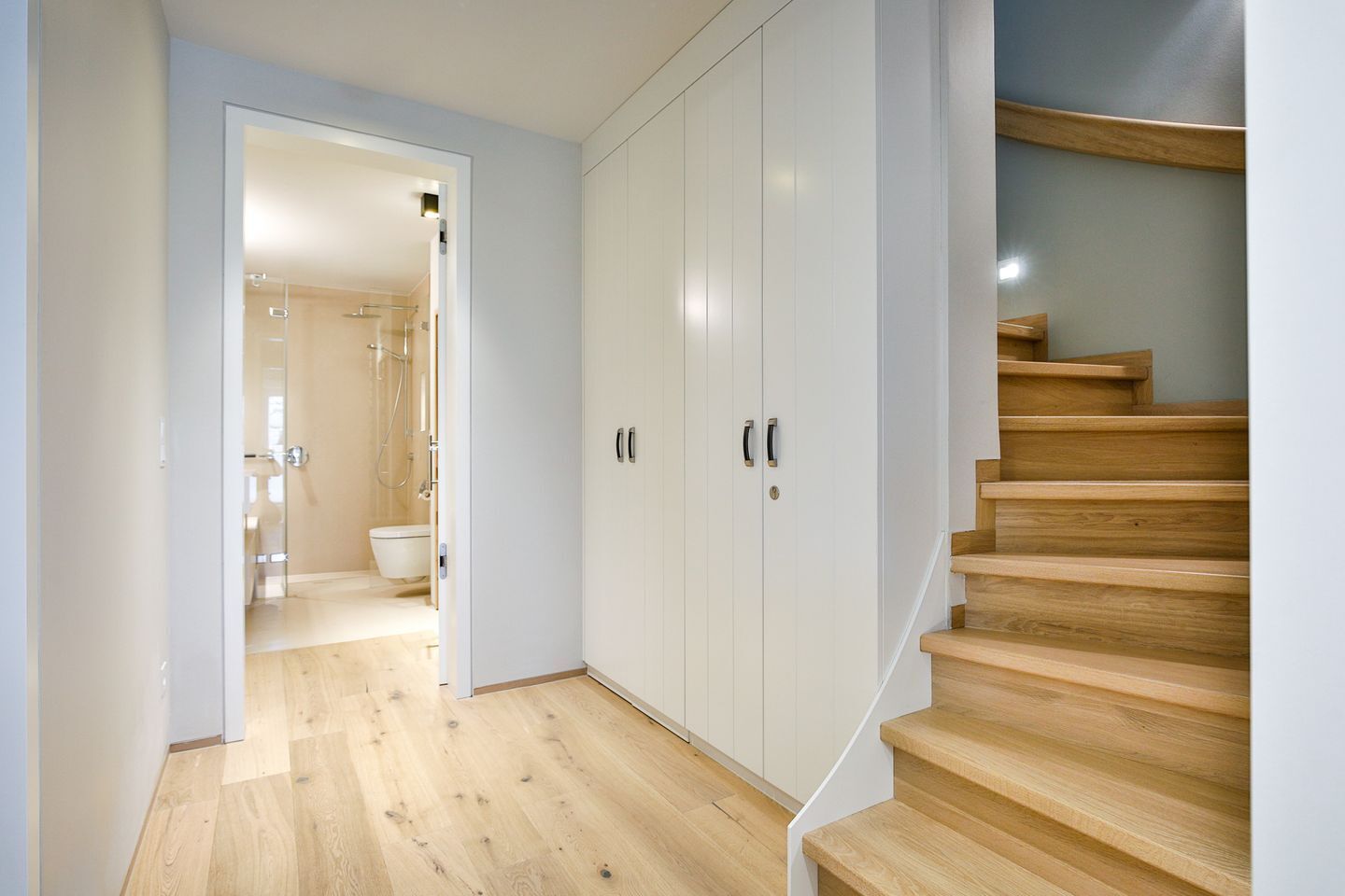 A hallway with wooden flooring leading to a bathroom with a shower and toilet. There is a staircase with wooden steps and a white wall on the right.