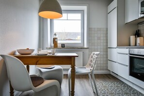 A modern kitchen with a dining area, featuring a wooden table, white chairs, a pendant light, and a view through a window. - Plyck-Hüs - Beautiful vacation home with a fenced-in yard, near the beach in (Hörnum (Sylt))