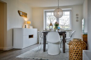 Un coin repas comprenant un meuble blanc, une table avec des chaises, un luminaire suspendu et une fenêtre donnant sur des arbres.
