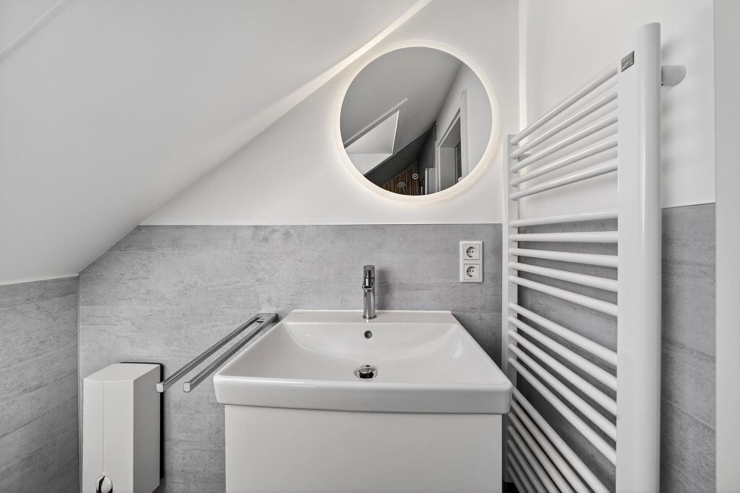 Un baño moderno con un lavamanos blanco, un espejo redondo y un perchero.