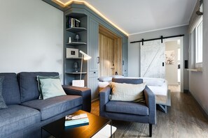 A modern living room with a sofa, armchair, and coffee table. There is a bookshelf with books and a lamp. A bed is visible in the background.