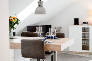 A dining area with a wooden table, four grey upholstered chairs, two wine glasses, and a vase with flowers. - Frisch Sanierte Wohnung in Ruhiger, Strandnaher Lage von Hörnum. - Berliner Ring (Hörnum (Sylt))