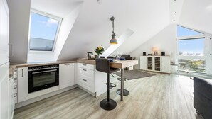 A modern kitchen with a dining area, white cabinetry, and a large window offering a view of the outdoors. - Frisch Sanierte Wohnung in Ruhiger, Strandnaher Lage von Hörnum. - Berliner Ring (Hörnum (Sylt))