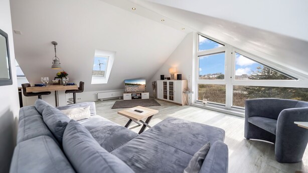 A modern loft apartment with a high ceiling, a dining area, a living room with a gray sofa, and a large window offering a view of the outdoors. - Frisch Sanierte Wohnung in Ruhiger, Strandnaher Lage von Hörnum. - Berliner Ring (Hörnum (Sylt))