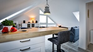 A modern kitchen with a wooden island, a dining area with a blue sofa, and a view of the city through large windows. - Frisch Sanierte Wohnung in Ruhiger, Strandnaher Lage von Hörnum. - Berliner Ring (Hörnum (Sylt))
