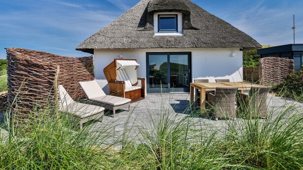 A thatched-roof house with a patio area, including a wooden table and two lounge chairs. - Beach house - Fantastic detached house under thatch - in the immediate vicinity of the beach. (Hörnum (Sylt))