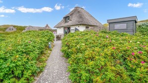A thatched-roof house with a stone pathway leading to it, surrounded by lush greenery and flowers. - Beach house - Fantastic detached house under thatch - in the immediate vicinity of the beach. (Hörnum (Sylt))