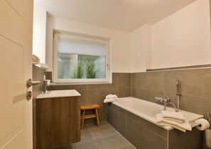 A modern bathroom with a bathtub, a wooden stool, and a window with blinds.