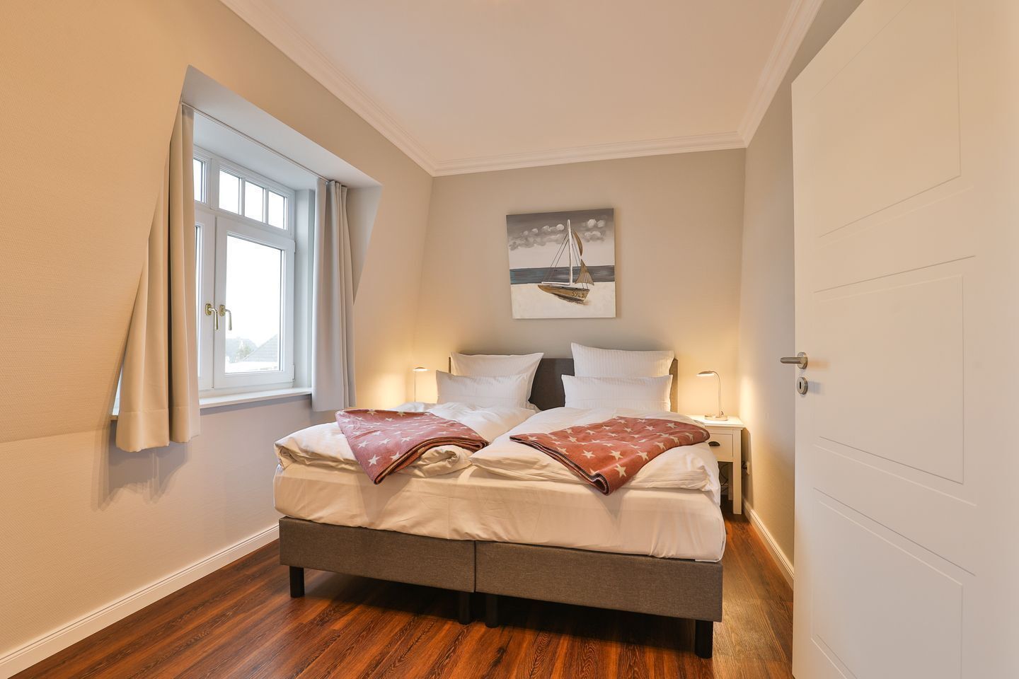 Ein Schlafzimmer mit einem Bett, einem Fenster mit Vorhängen, einem Nachttisch mit Lampe und einem Bild an der Wand.