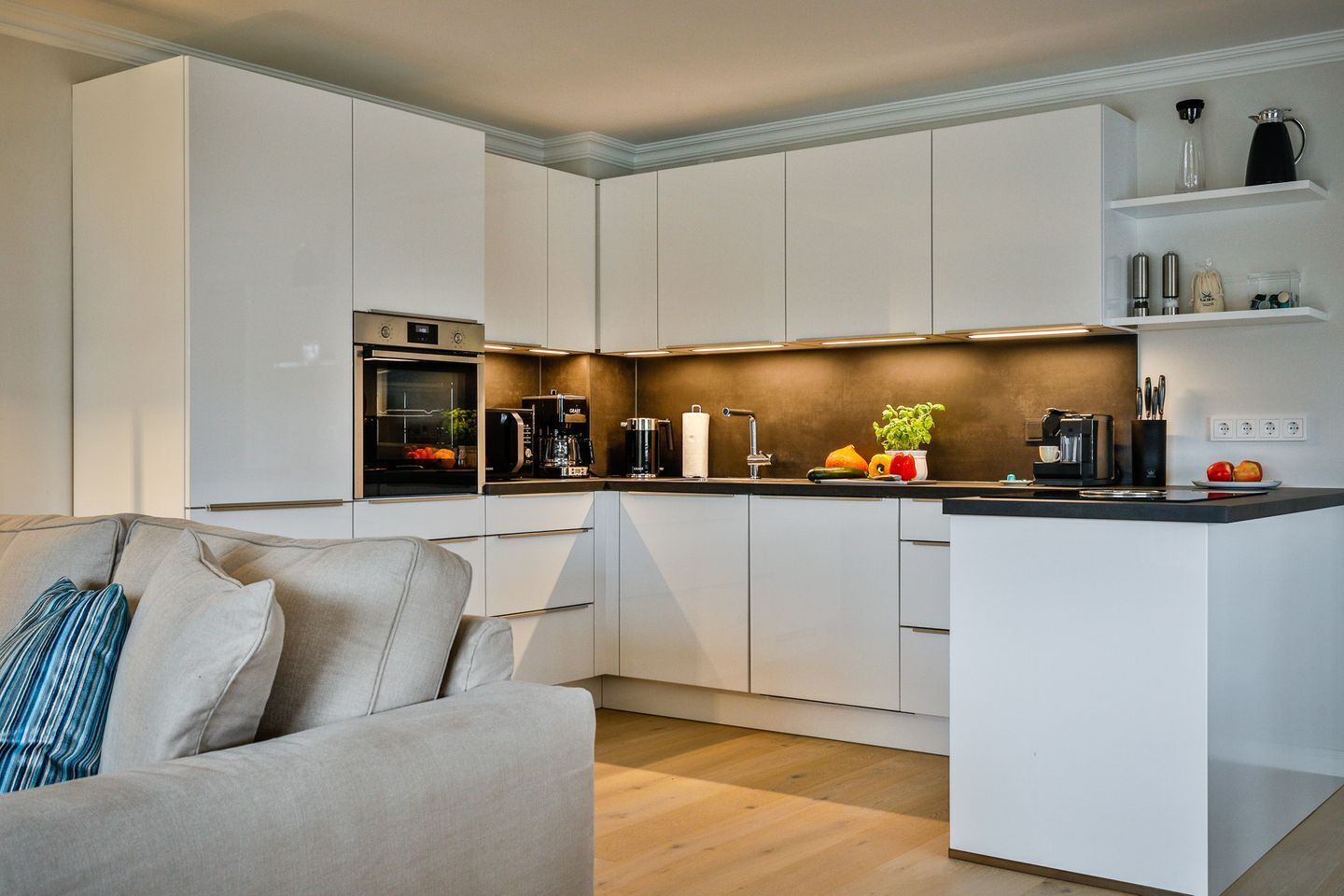 A modern kitchen with white cabinetry, a built-in oven, a coffee station, and a small island with fruits and vegetables.