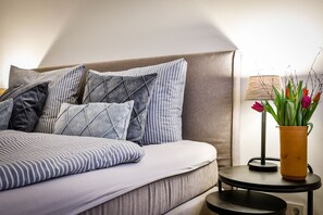 A neatly made bed with striped bedding and geometric pillows, a small round table with a lamp and vase of flowers, and a plain wall in the background. - Lornsendüne - Modern apartment in an absolutely beachfront location in Westerland with (Sylt)