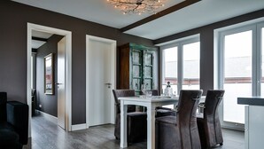 Een eetruimte met een witte tafel en donkere stoelen, een houten kast en grote ramen.