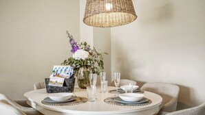 A round dining table with a white tablecloth, surrounded by beige upholstered chairs. A wicker pendant light hangs above. A vase with white and purple flowers and a basket with a card and pens are on the table.