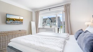 A bedroom with a bed, a wicker chair, a window with curtains, and a painting on the wall.