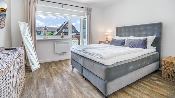A modern bedroom with a large bed, a window with curtains, a radiator, and a wicker side table.