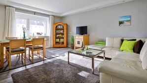 Ein Wohnzimmer mit einer Couch, einem Couchtisch und einem Fernseher. Es gibt einen Essbereich mit Stühlen und einem Tisch. Ein Bücherregal mit dem Schriftzug „HOME“ ist sichtbar.