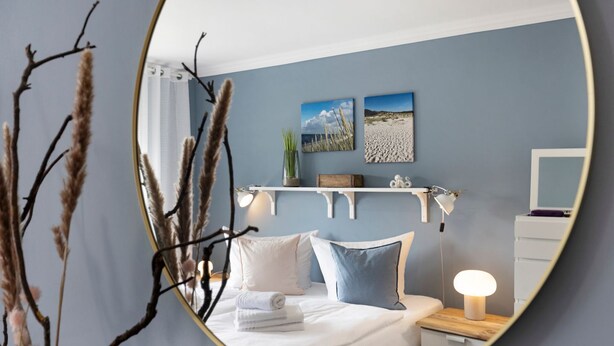A bedroom with a bed, bedside table, lamp, and wall-mounted shelf with decorative items. - Njørd - Haus Möwe, Deutschland (Hörnum (Sylt))