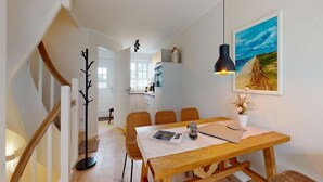 A dining area with a wooden table, wicker chairs, a black pendant light, and a framed painting of a landscape. - Ellis beach house Sylt (Hörnum (Sylt))